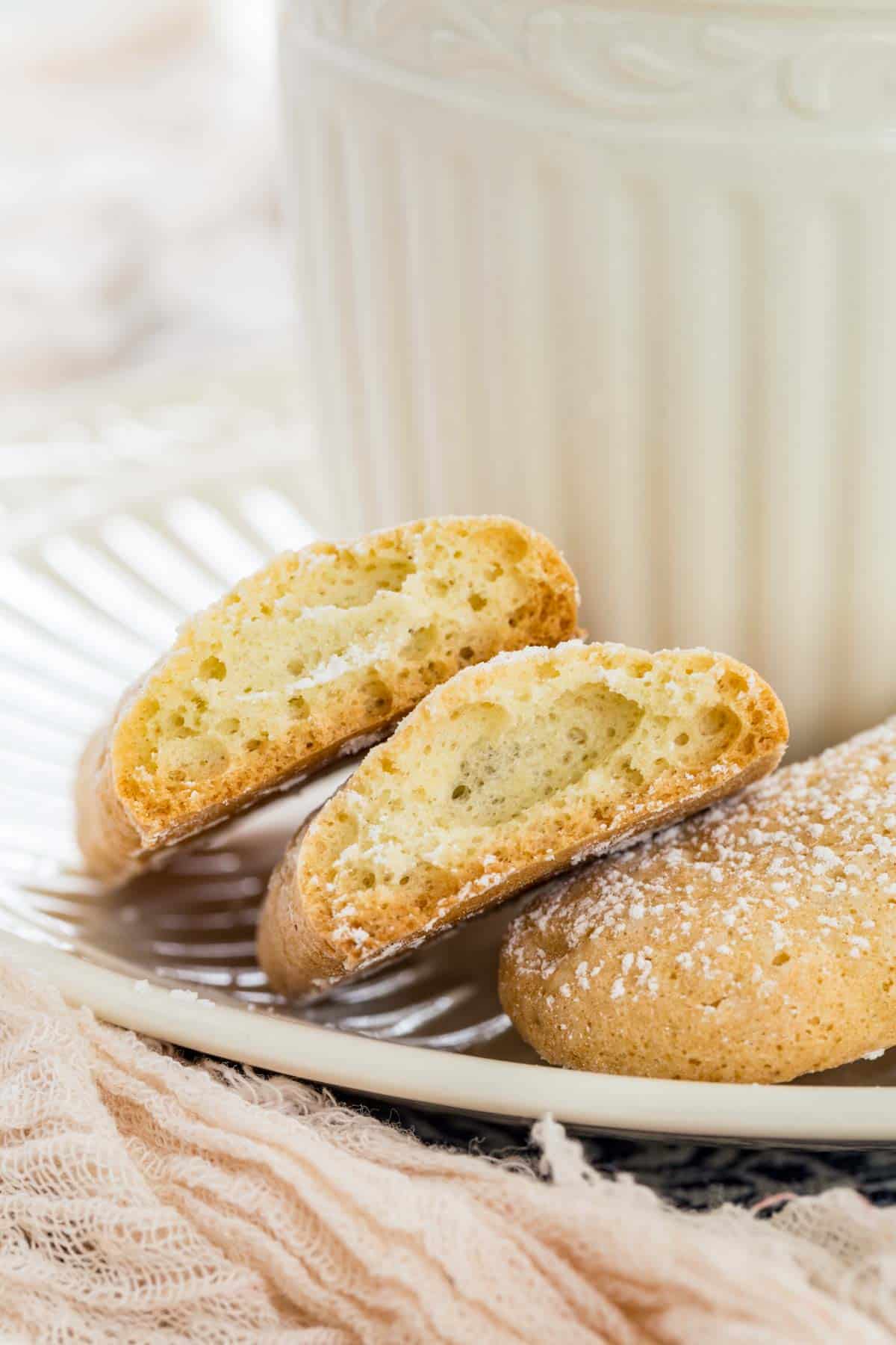 Close up of two halves of a gluten free ladyfinger cookie on a saucer next to a cup of coffee.