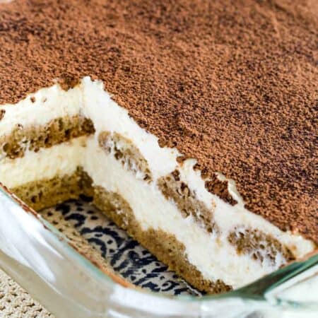 Gluten-free tiramisu dusted with cocoa powder inside a glass baking dish, with two servings missing.