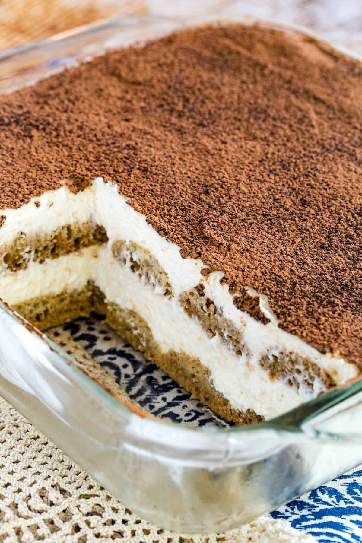 Gluten-free tiramisu dusted with cocoa powder inside a glass baking dish, with two servings missing.