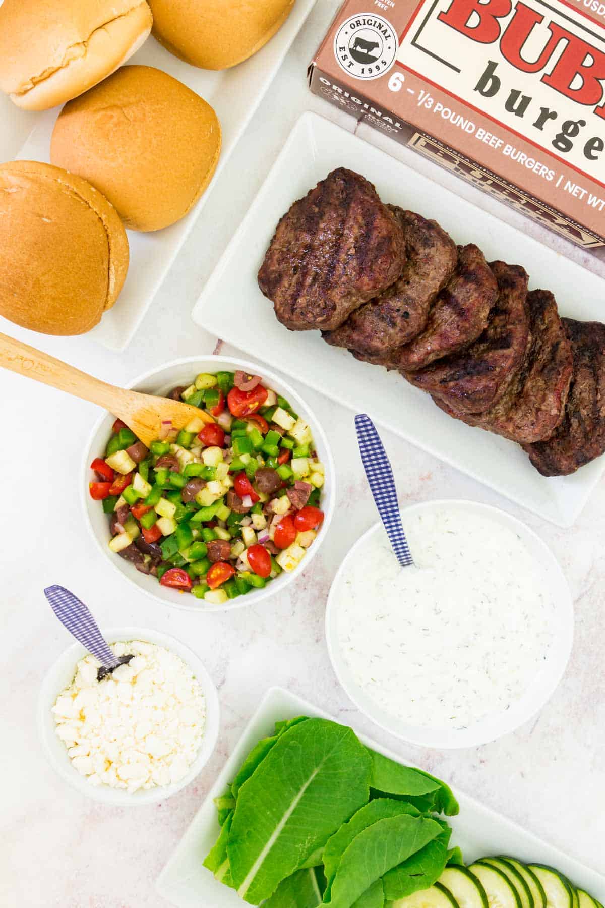Grilled burgers and buns on platters, a box of BUBBA burgers, and bowls of Greek burger toppings.