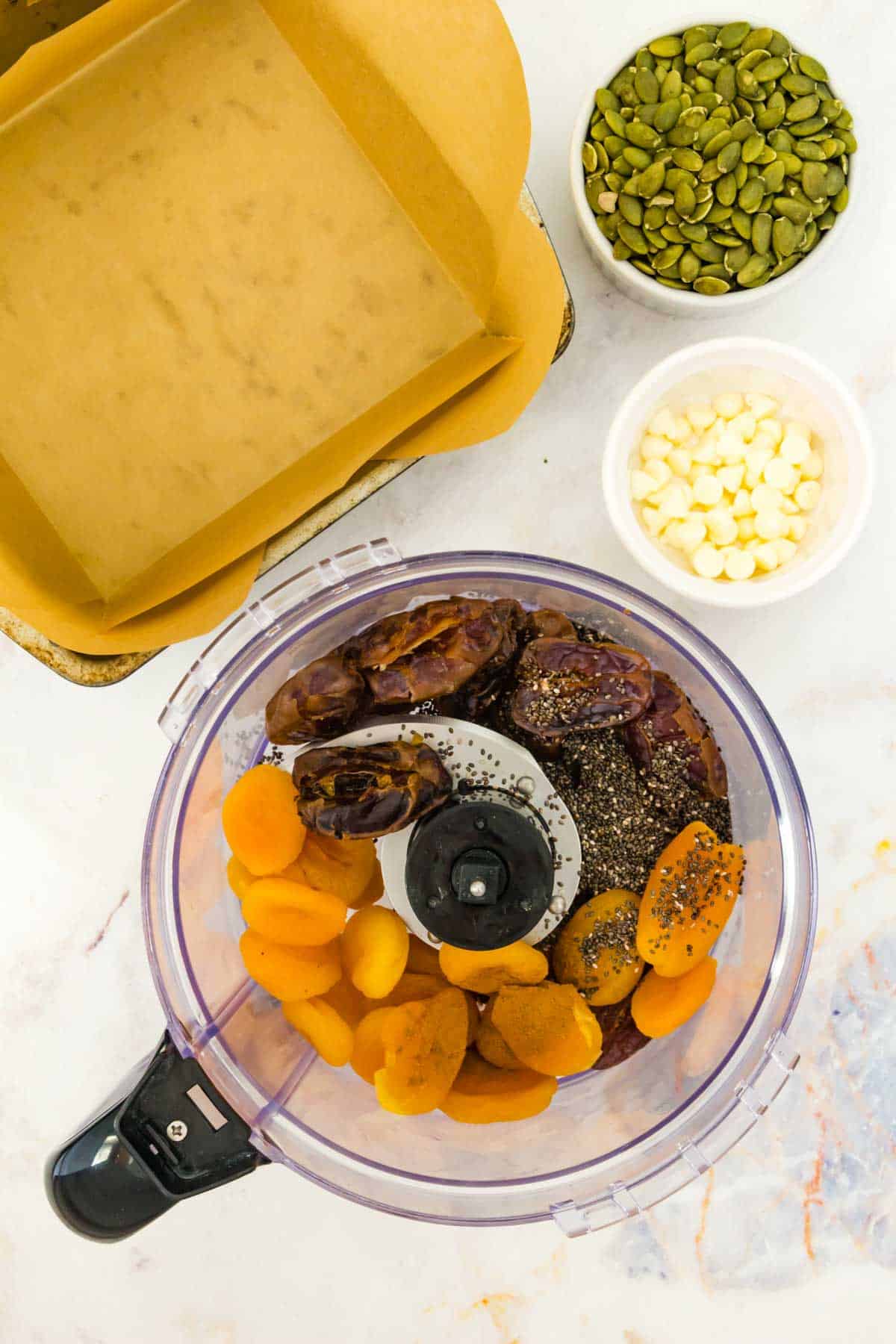 dried apricots, dates, and chia seeds in a food processor next to bowls of other ingredients.