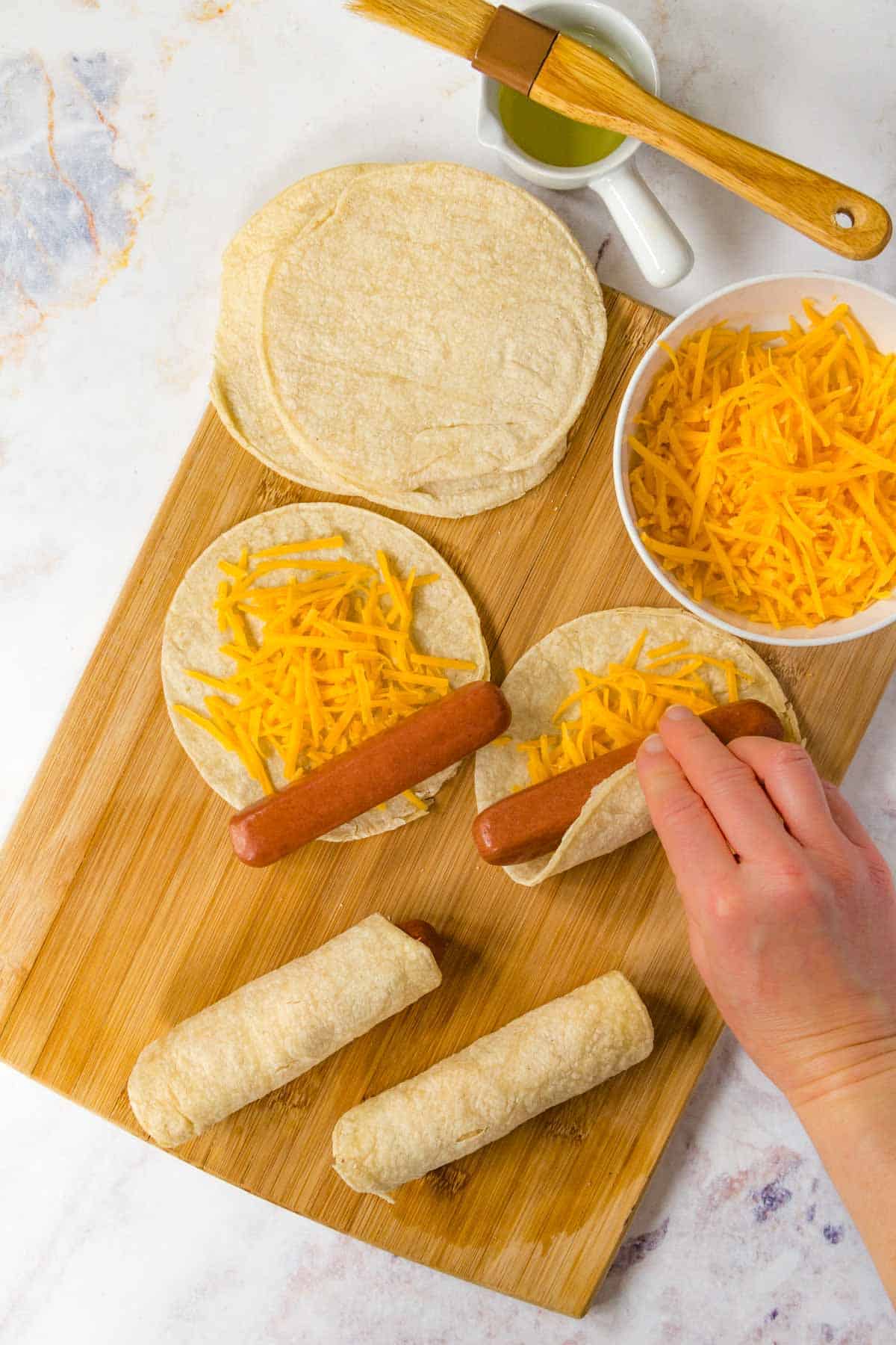 Hot dogs being rolled up in corn tortillas with shredded cheddar cheese on a wooden cutting board.