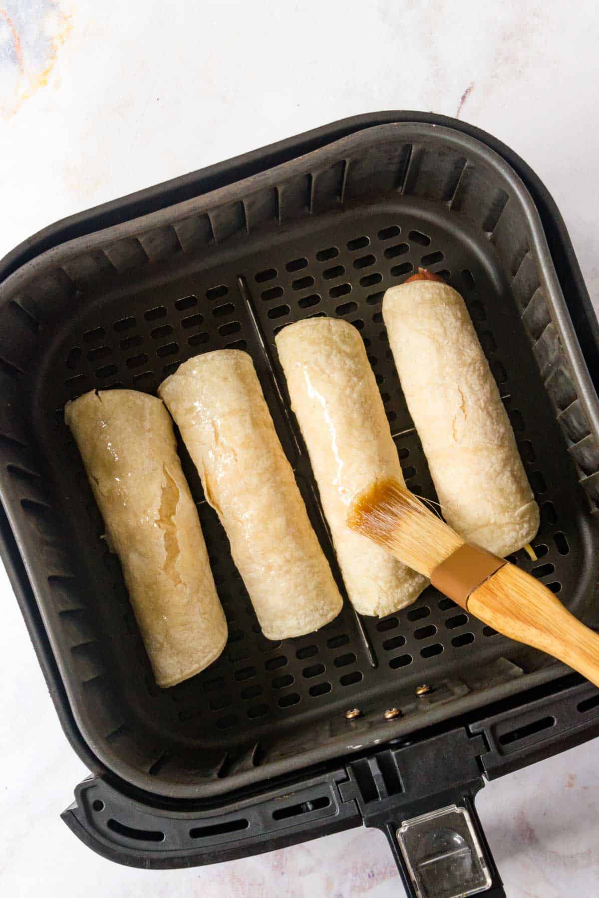 brushing oil on uncooked hot dogs wrapped in tortillas in an air fryer basket.