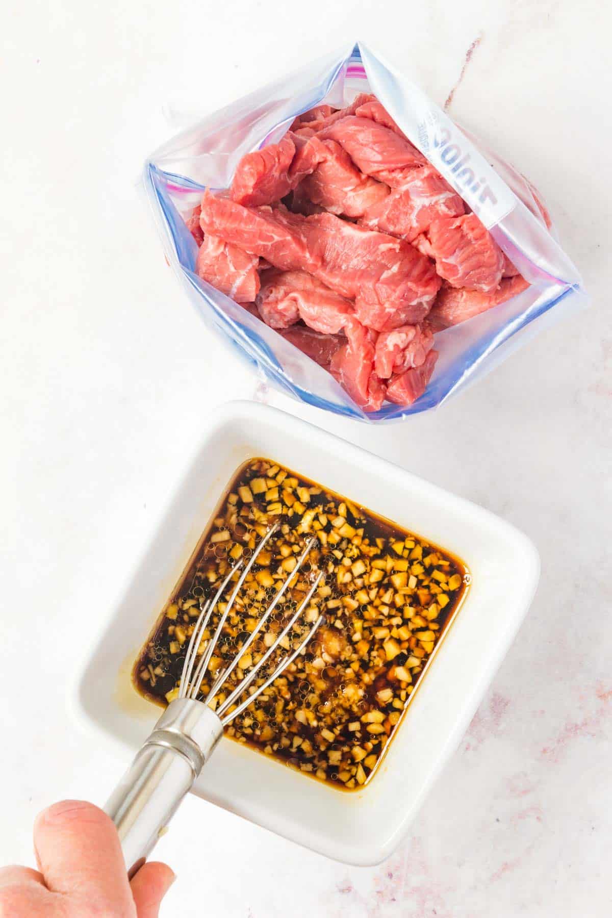 Whisking the marinade in a bowl next to a ziploc bag of sliced beef.