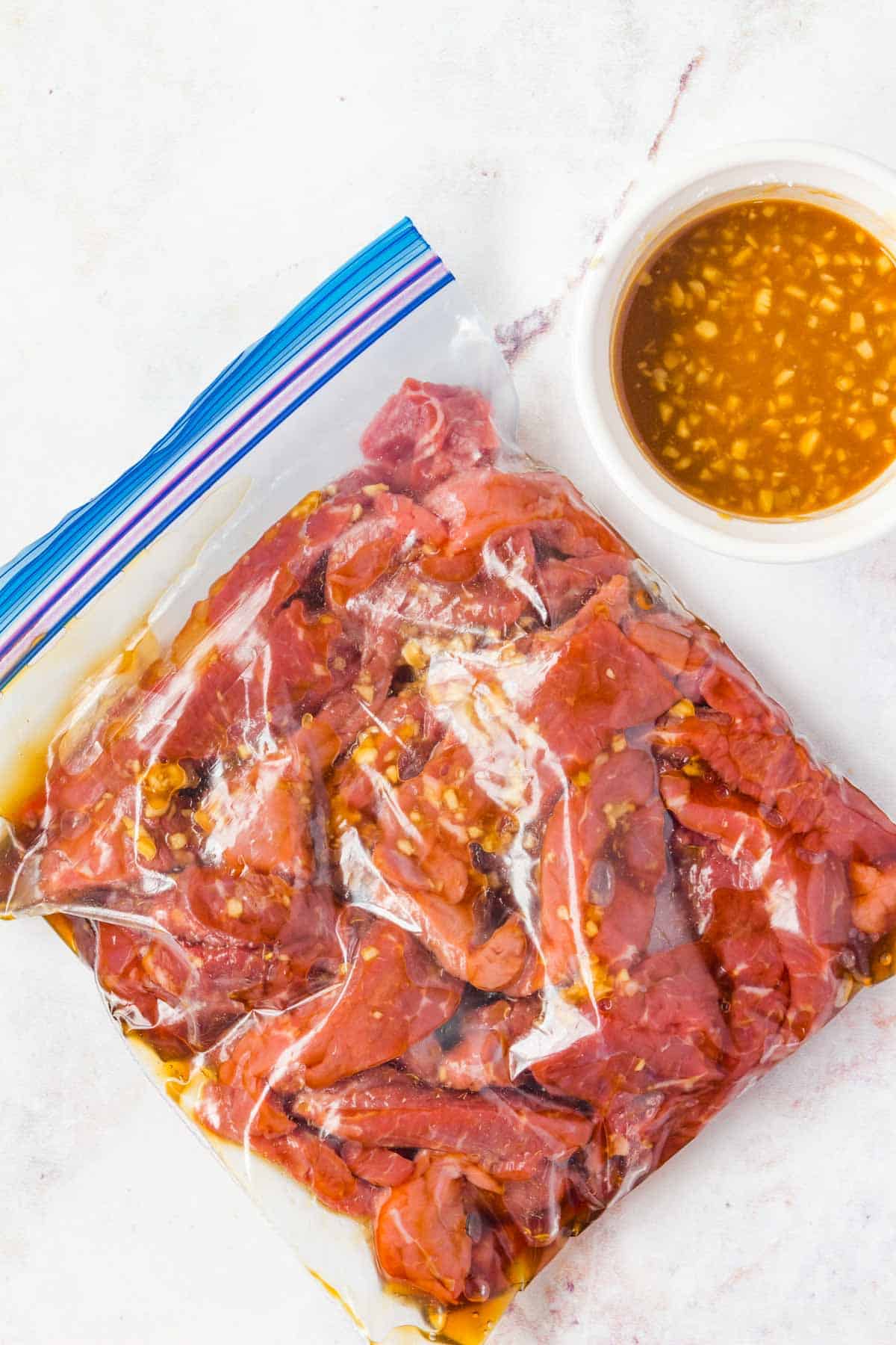 A plastic storage bag with marinating beef slices and small bowl of stir fry sauce.