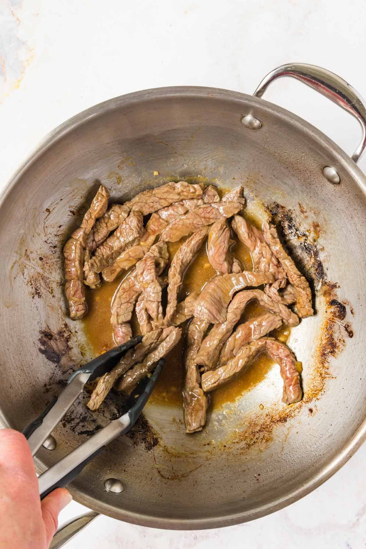 Browning the beef slices in a wok and turning them with tongs.