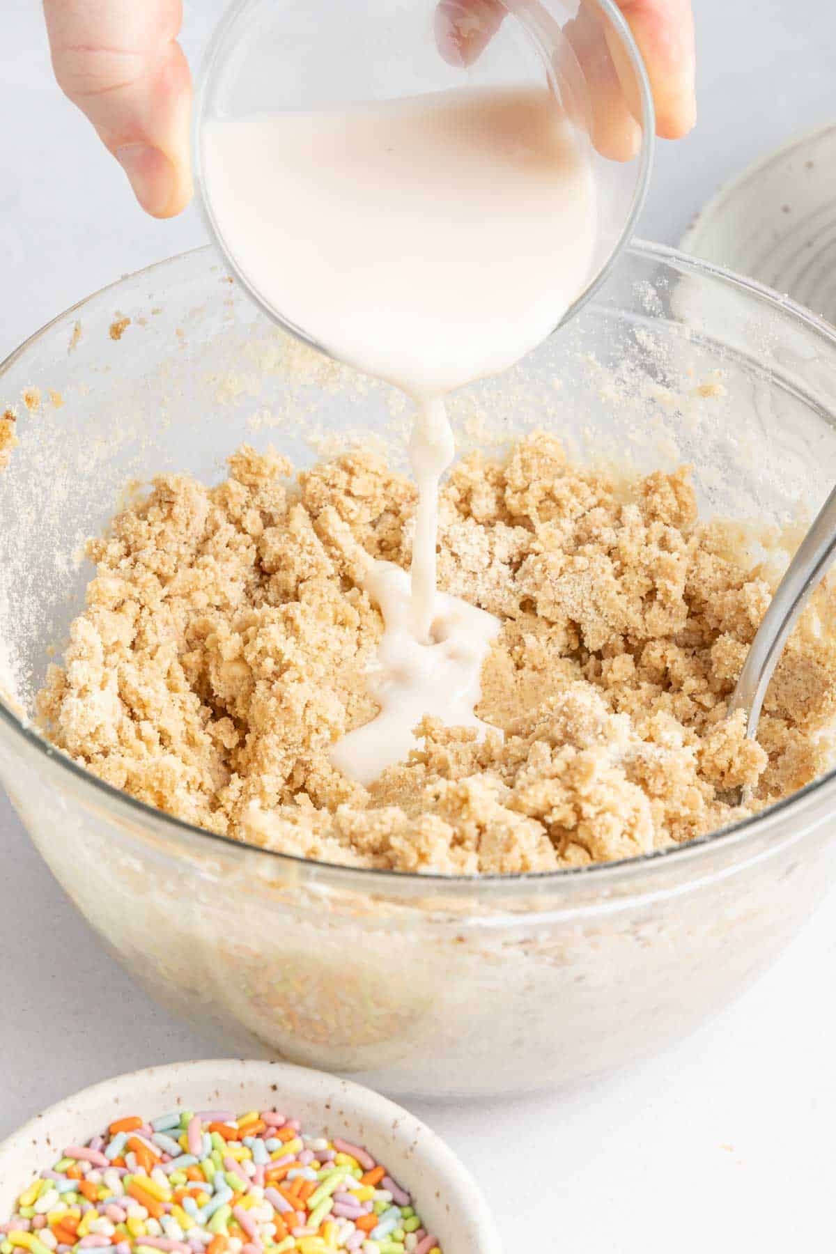 A hand pours milk into a glass bowl of mixed cookie dough.