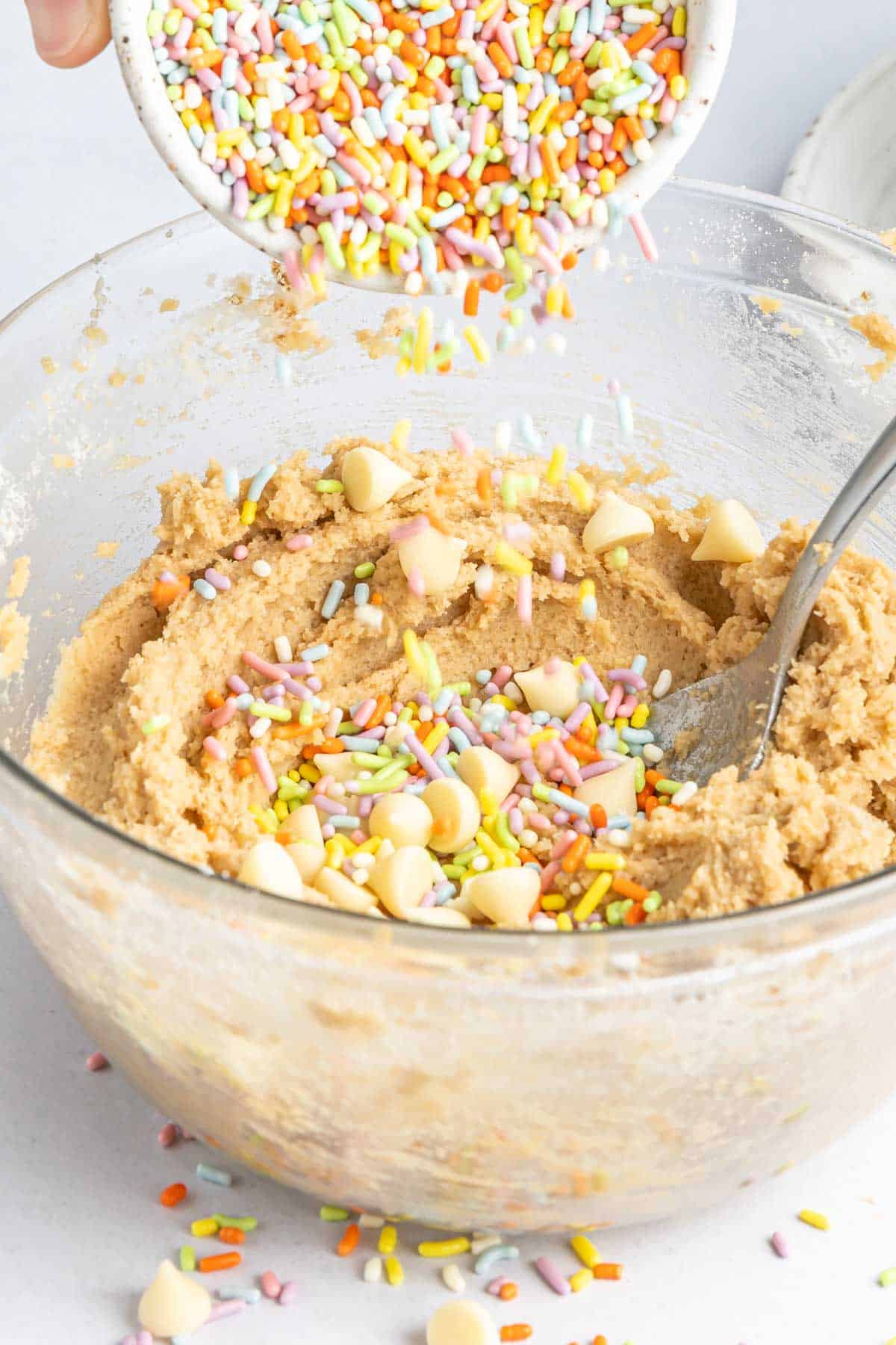 Sprinkles are poured into a glass mixing bowl full of cookie dough mixed with white chocolate chips..