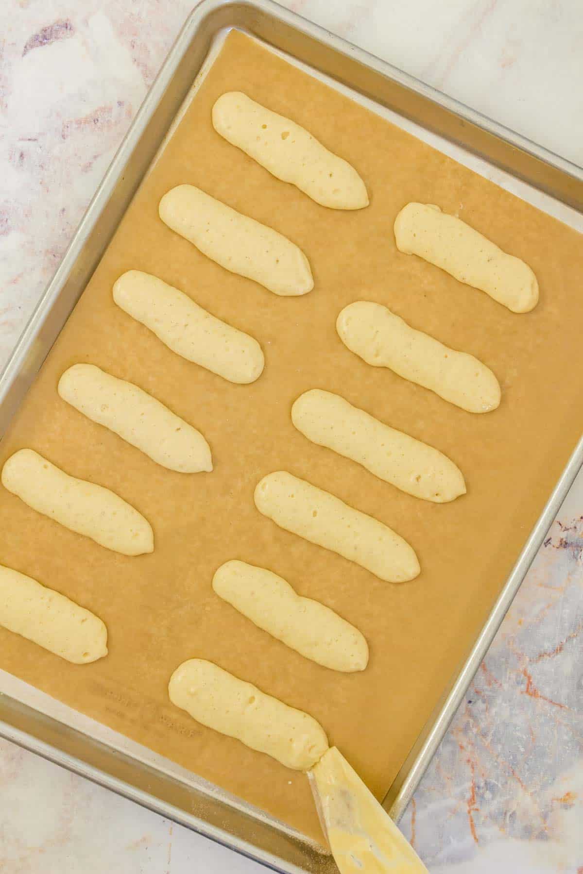 A piping bag is used to pipe lines of ladyfinger batter onto a parchment lined baking sheet.