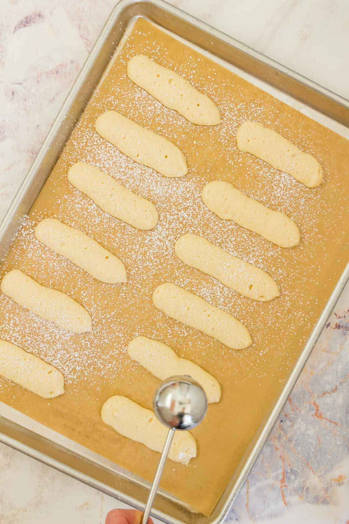 Unbaked ladyfingers are dusted with powdered sugar on a parchment lined baking sheet.