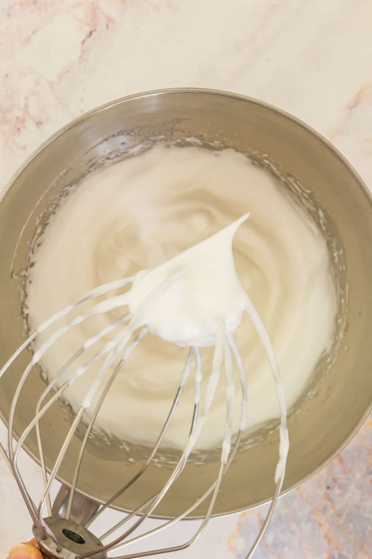A whisk with a meringue peak held up in front of whipped egg whites and sugar in a mixing bowl.