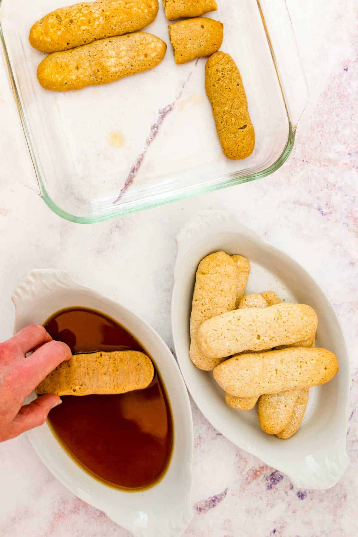 A hand dips a gluten-free ladyfinger into a mixture of espresso and rum, next to a bowl of ladyfingers and a baking dish with dipped ladyfingers.