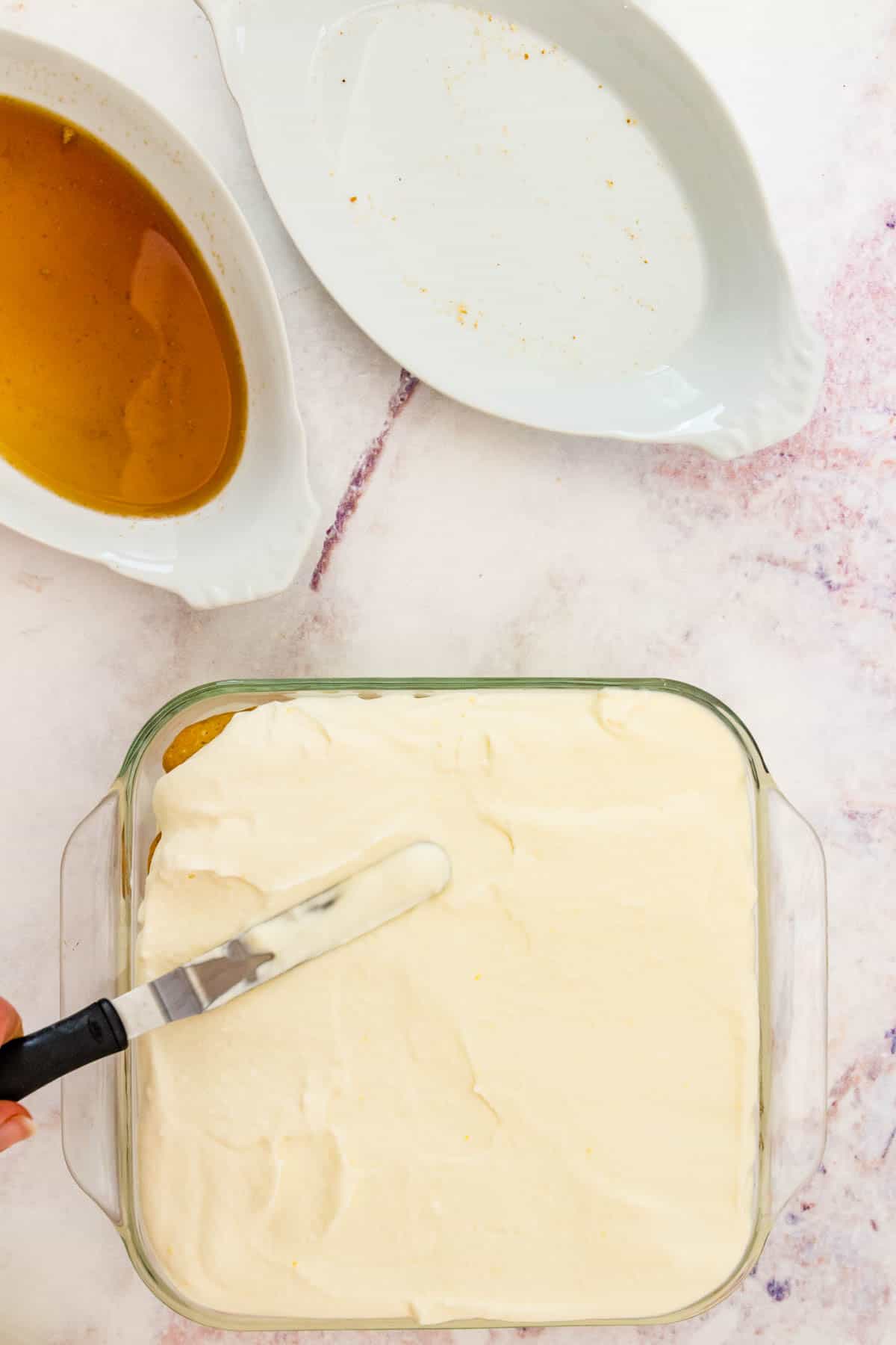 A final layer of mascarpone is spread over the other tiramisu layers in a glass baking dish.