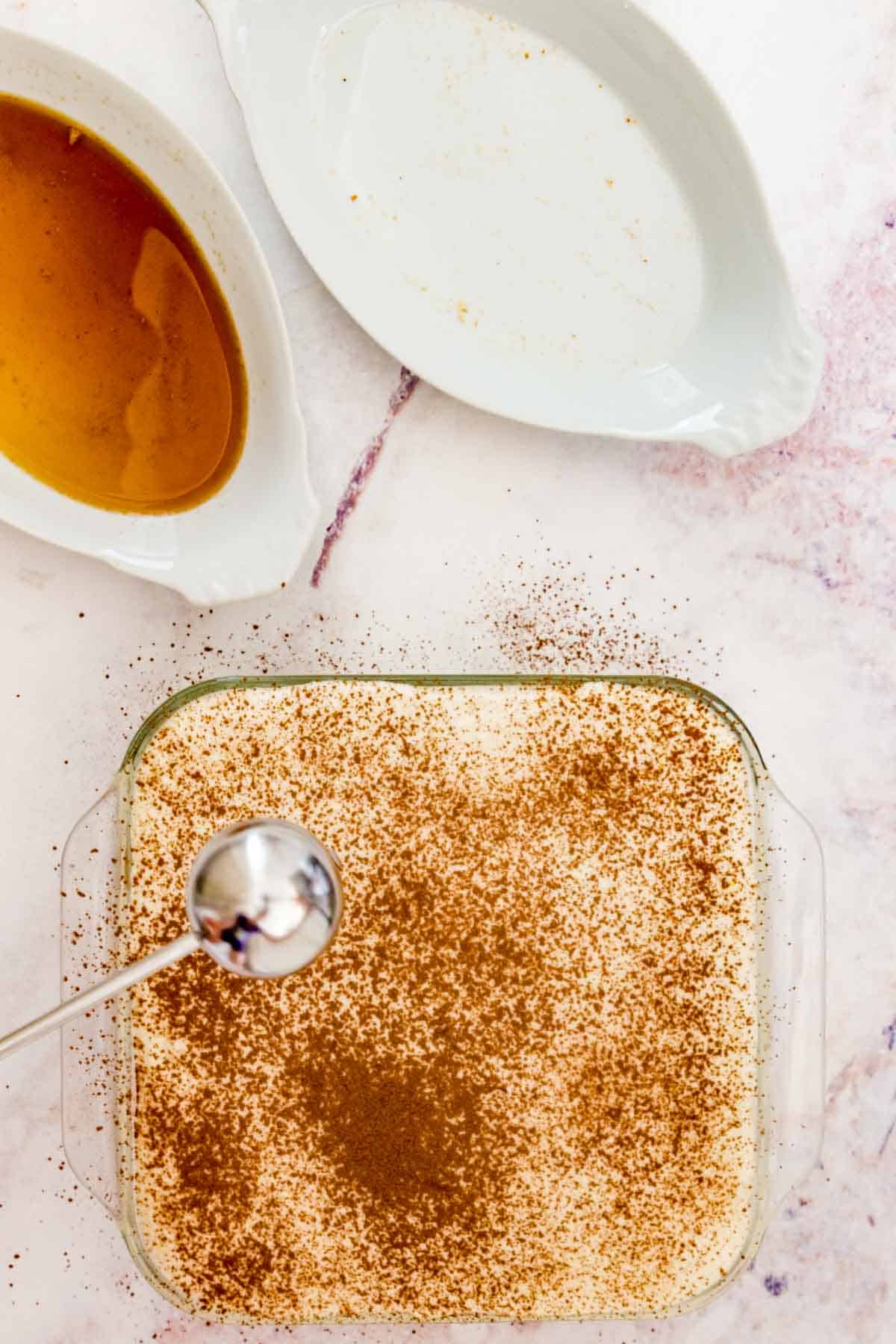 Cocoa powder is dusted over top of a finished tiramisu in a glass baking dish.