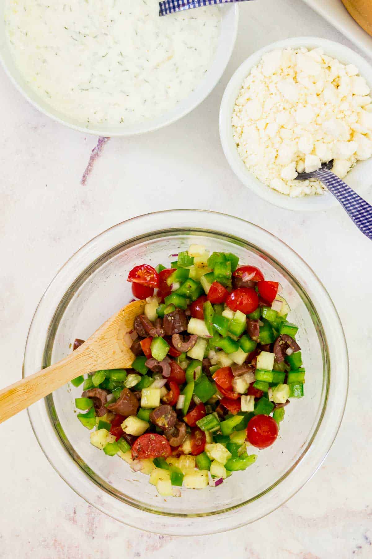Greek salad topping mixed in a bowl with bowls of tzatziki and feta next to it.