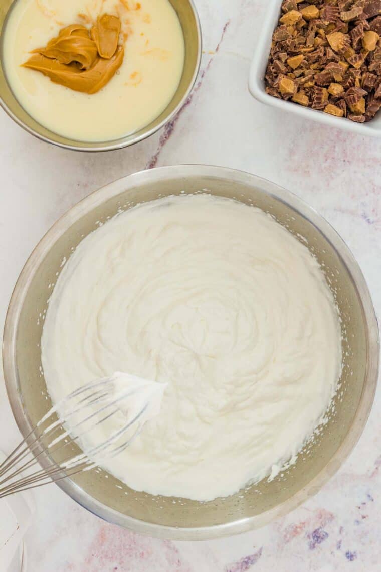 Whipped cream in a metal mixing bowl next to bowls of condensed milk with peanut butter and Mini Reese's Cups.