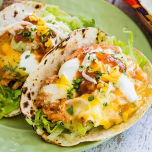 Two egg tacos on charred tortillas topped with cheese, sour cream, and other toppings on a small green plate.