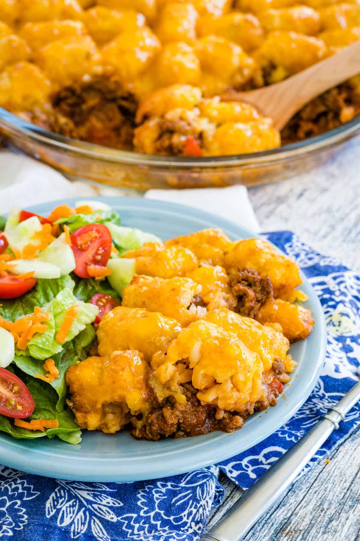 A serving of sloppy joe tater tot casserole on a plate next to a green salad, with the rest of the casserole in the background.