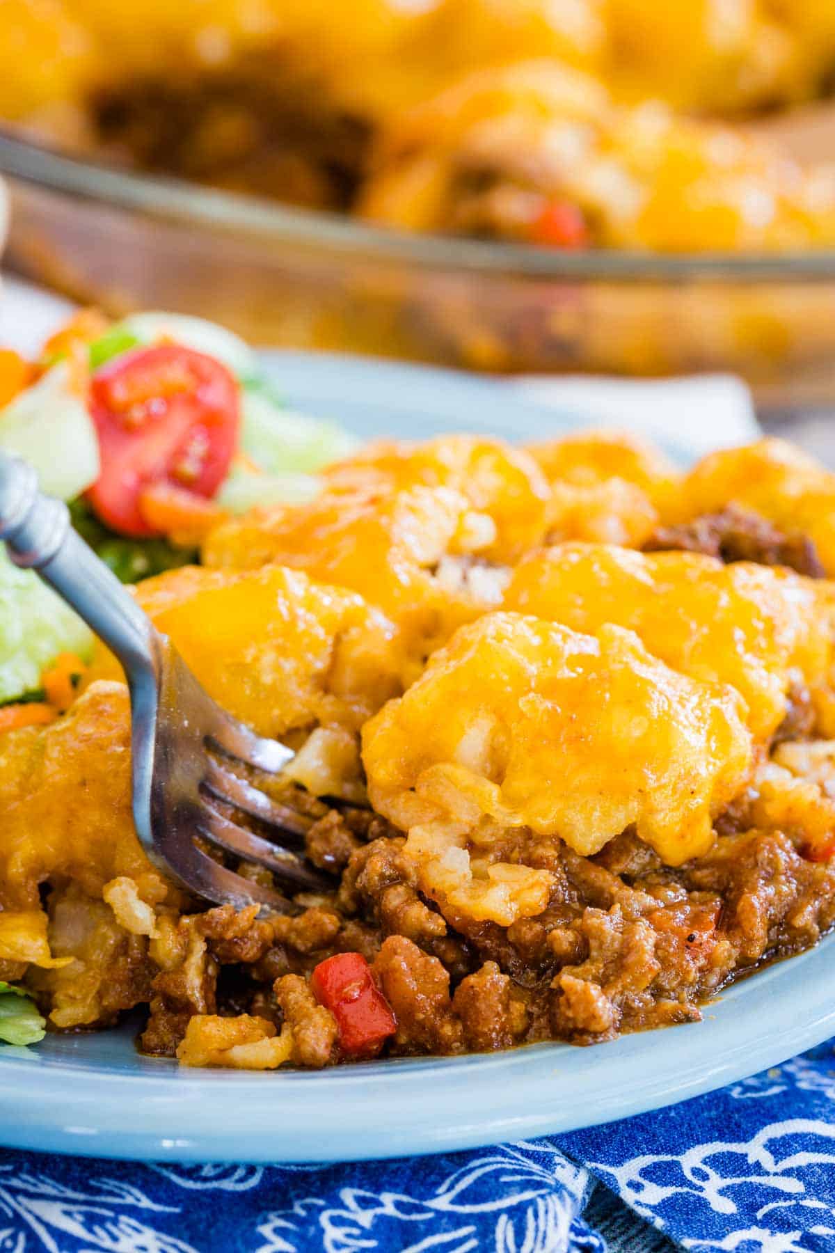 A fork stuck into a serving of sloppy joe tater tot casserole on a plate next to a green salad.