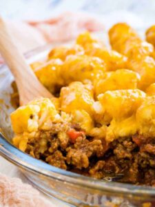 A wooden spoon scoops a serving of sloppy joe tater tot casserole from a baking dish.