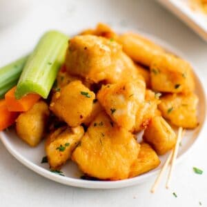 A small white dish of buffalo chicken bites with two toothpicks and celery and carrot sticks.