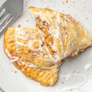 An apple turnover broken in half on a plate and drizzled with glaze.
