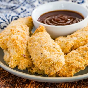 Breaded baked chicken tenders on a dish with a small bowl of barbecue sauce.