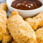 A closeup of baked chicken tenders on a dish with a bowl of barbecue sauce.