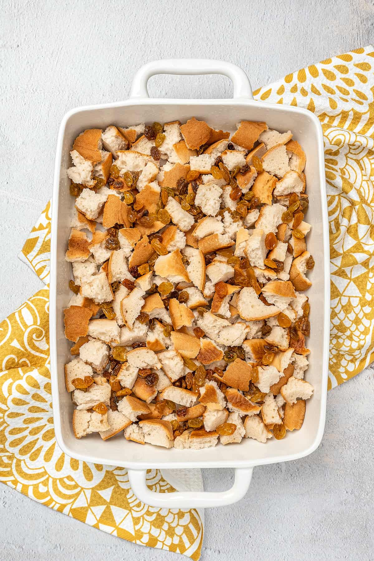 Cubed bagels and raisins spread into the bottom of a large rectangular baking dish.