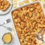 Overhead view of bread pudding in a baking dish next to a serving of bread pudding on a plate, next to a pourer with bourbon sauce.