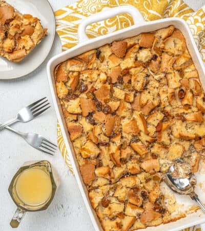 Overhead view of bread pudding in a baking dish next to a serving of bread pudding on a plate, next to a pourer with bourbon sauce.