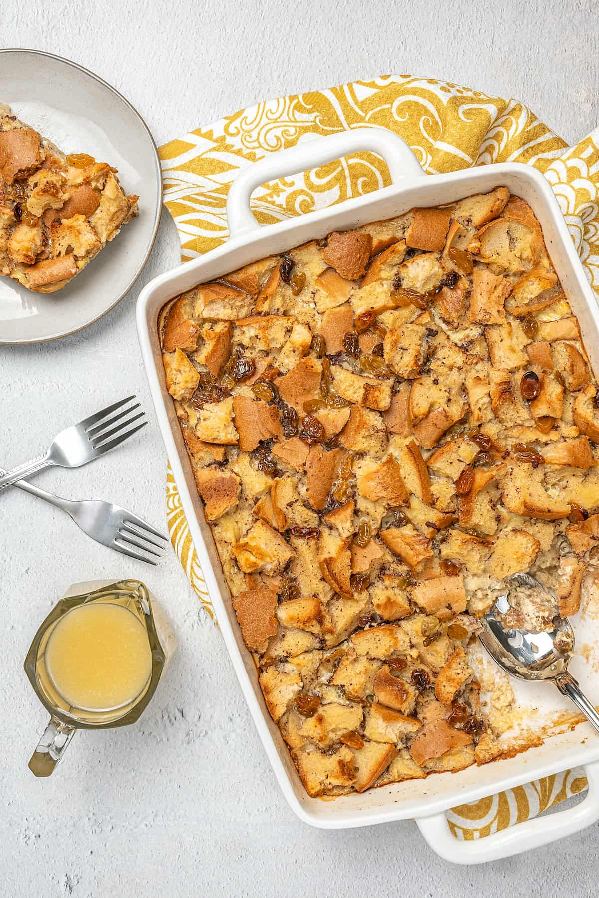 Overhead view of bread pudding in a baking dish next to a serving of bread pudding on a plate, next to a pourer with bourbon sauce.