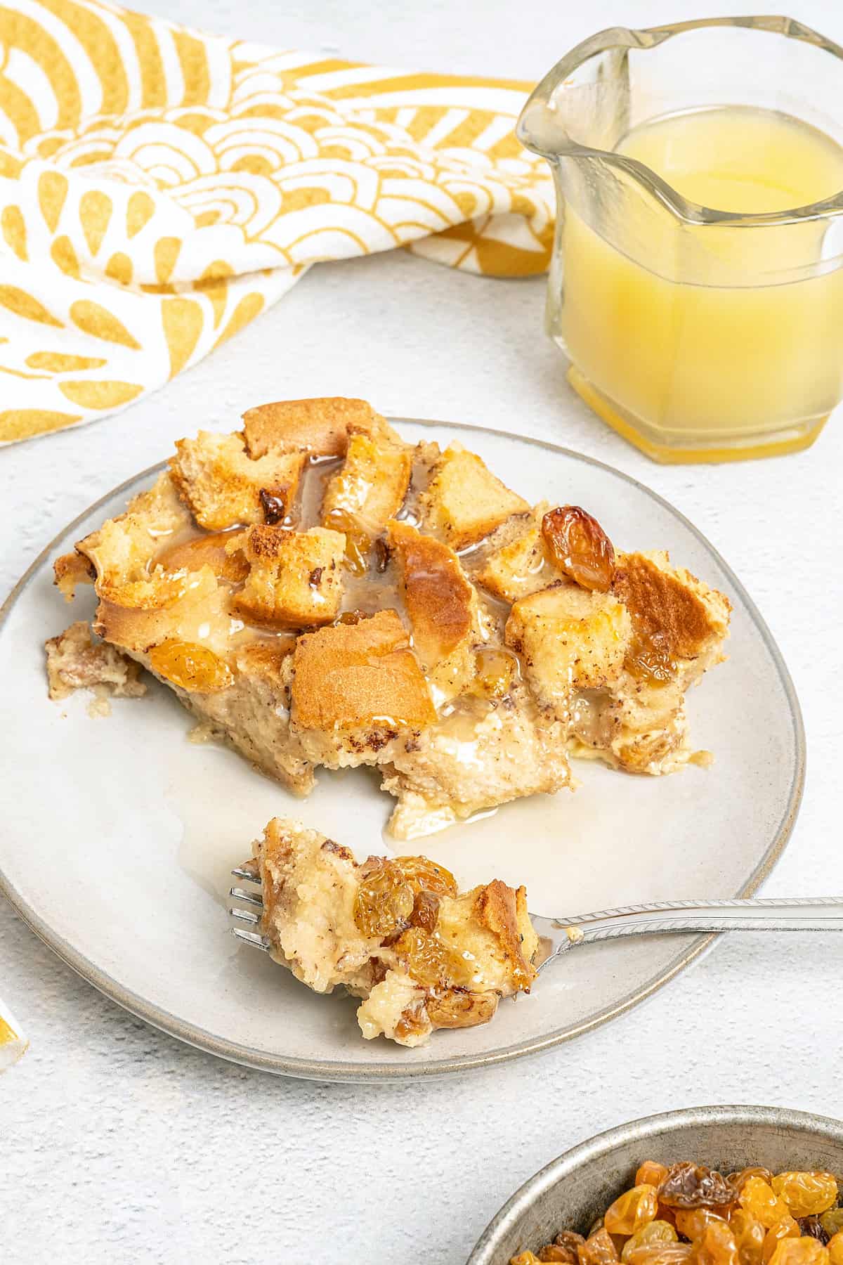 A slice of gluten-free bread pudding on a plate with a fork, with a pourer full of bourbon sauce in the background.
