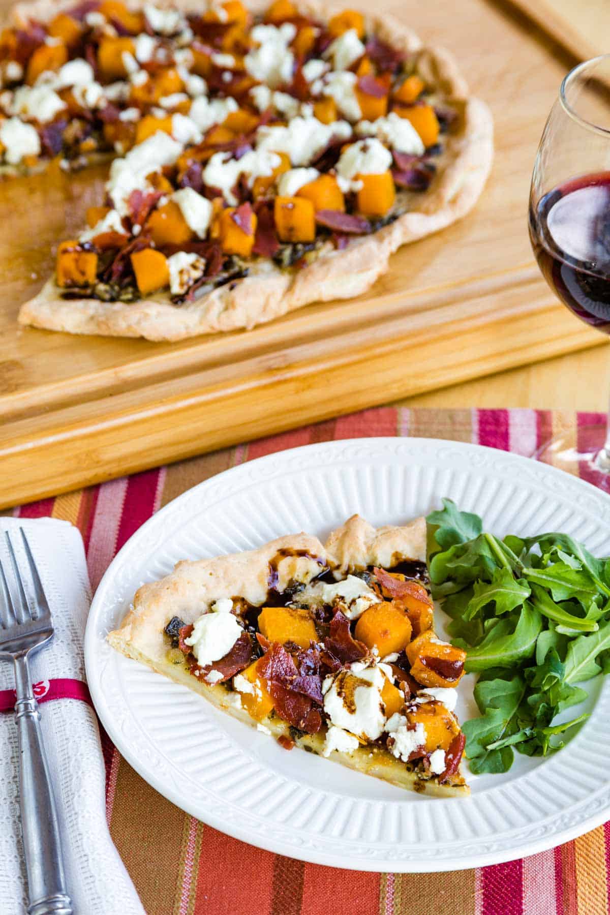 A plate of arugula and a slice of butternut squash pizza on a striped placement in front of a cutting board with the rest of the pizza.