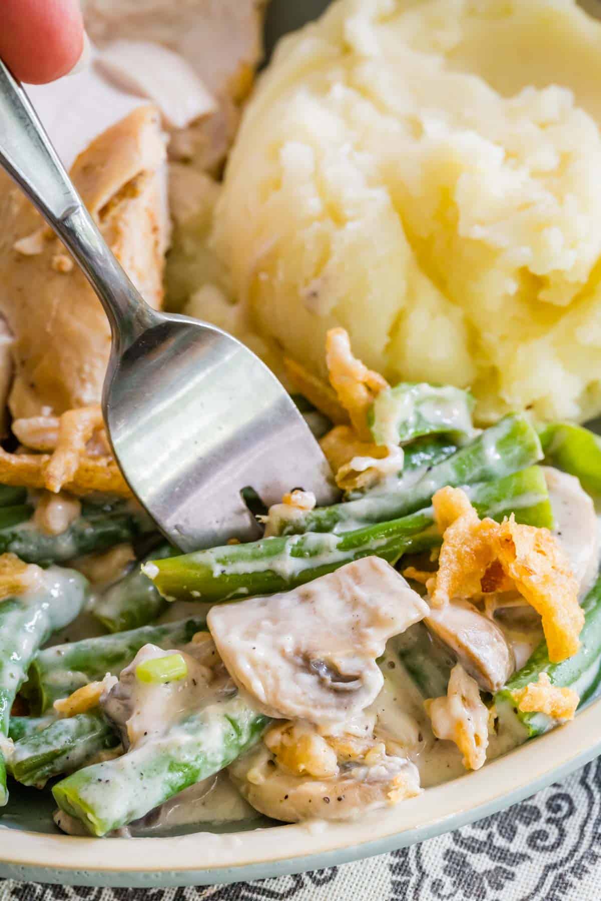A fork stuck into a serving of gluten-free green bean casserole on a plate next to mashed potatoes and slices of turkey.