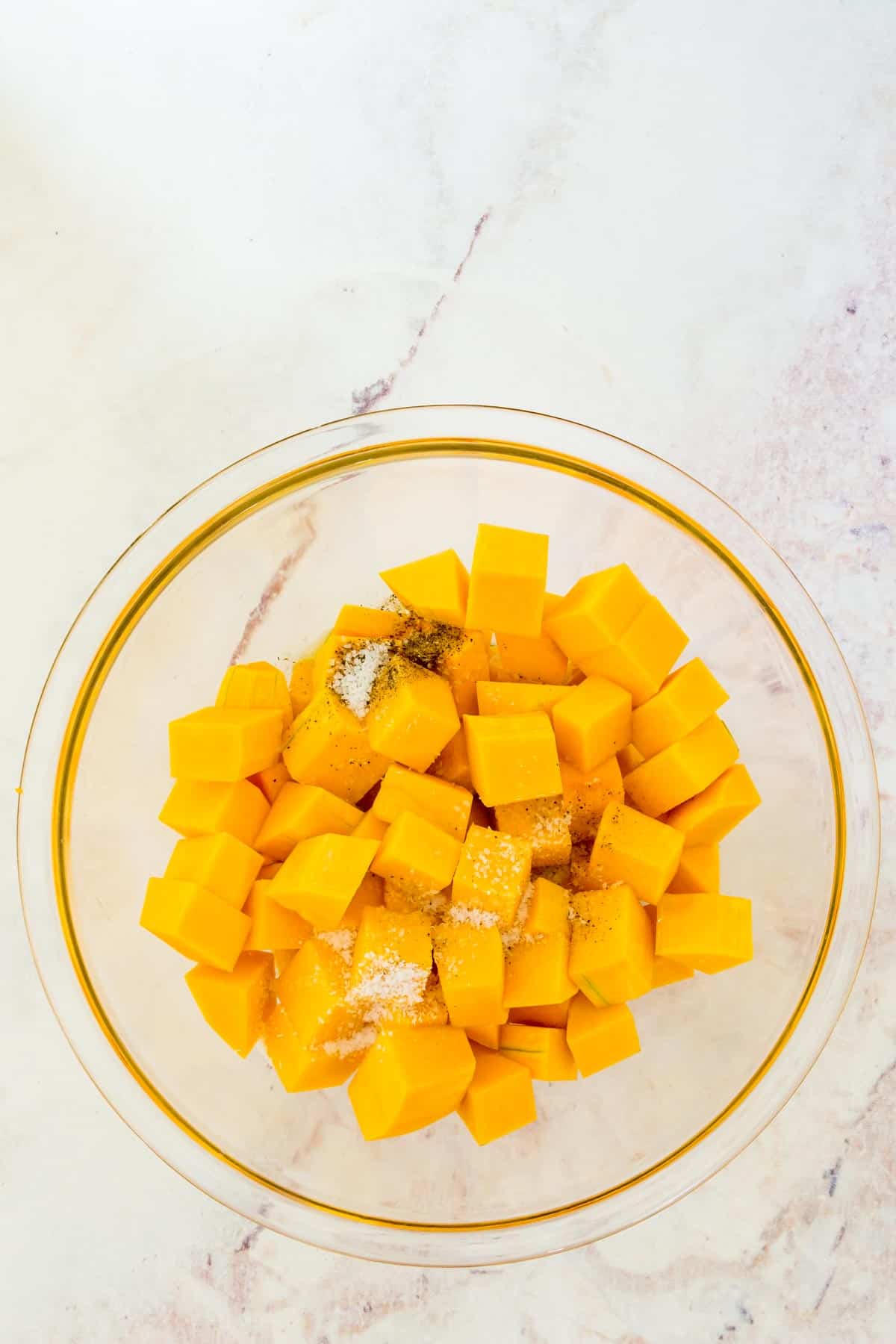 Cubed butternut squash combined with seasoning in a glass bowl.