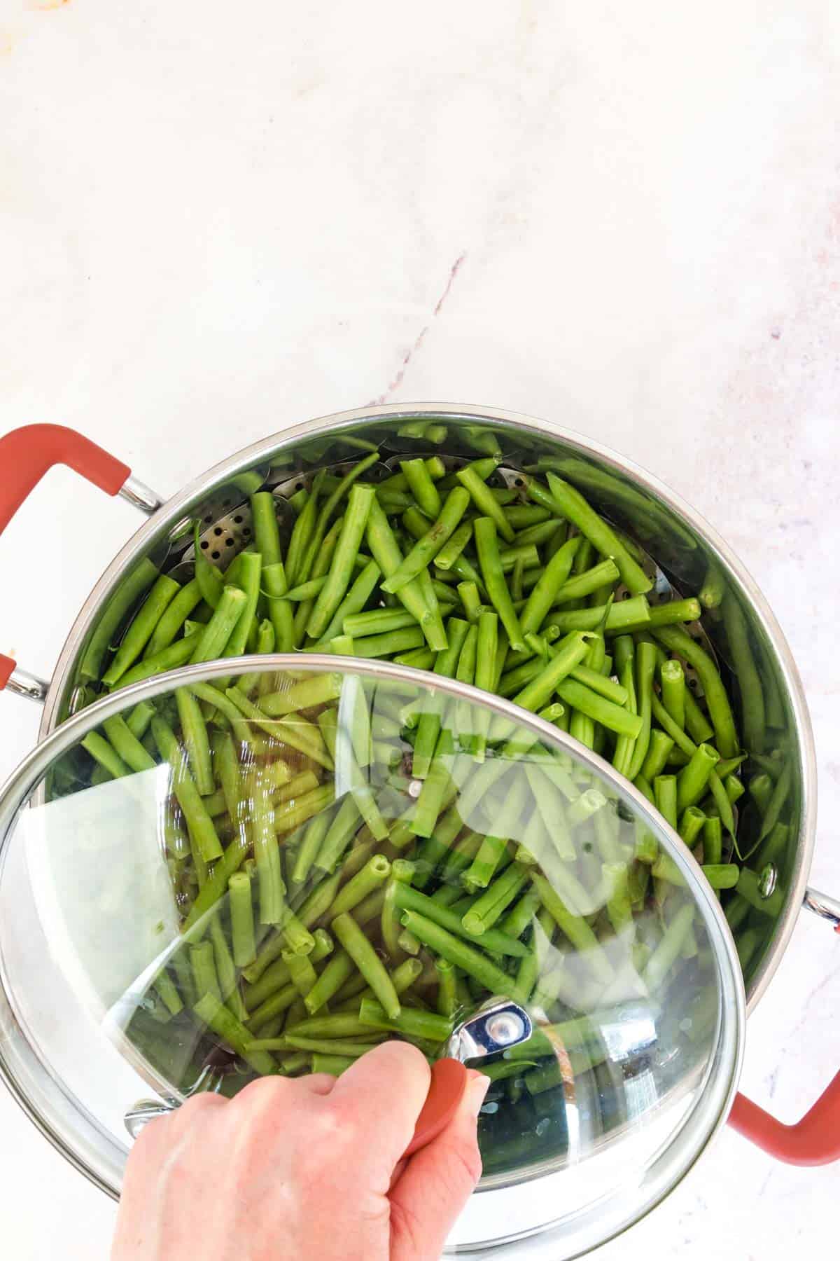 A hand holds the glass lid over a pot filled with green beans.