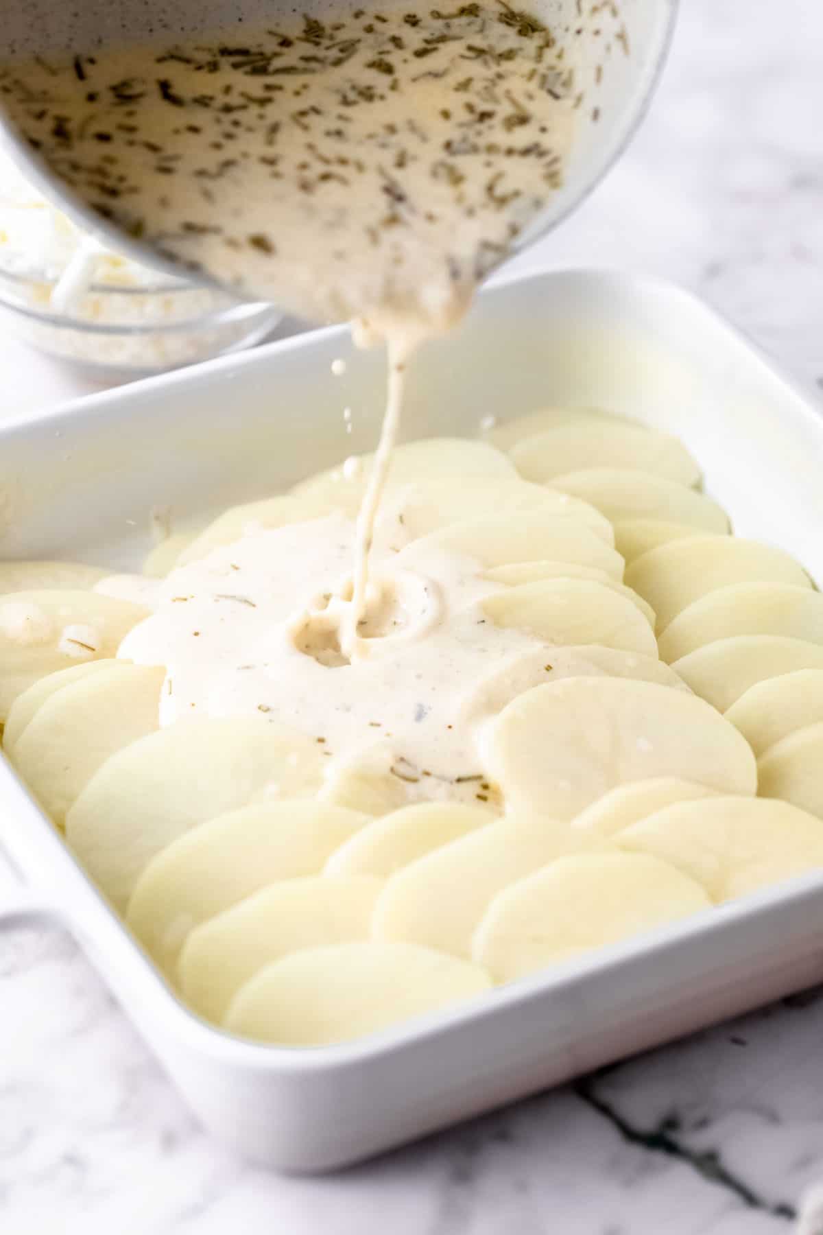 Cream sauce is poured from a saucepan over top potato slices in a baking dish.