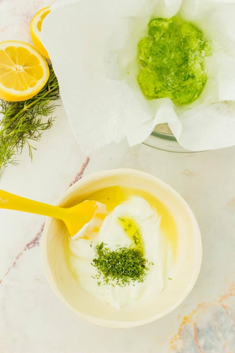 Greek yogurt, dill, lemon juice and some of the tzatziki ingredients in a bowl with a yellow spatula.