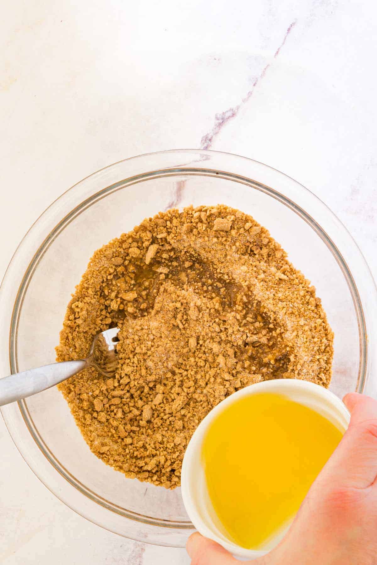 Melted butter is poured into a glass mixing bowl with graham cracker crust ingredients.