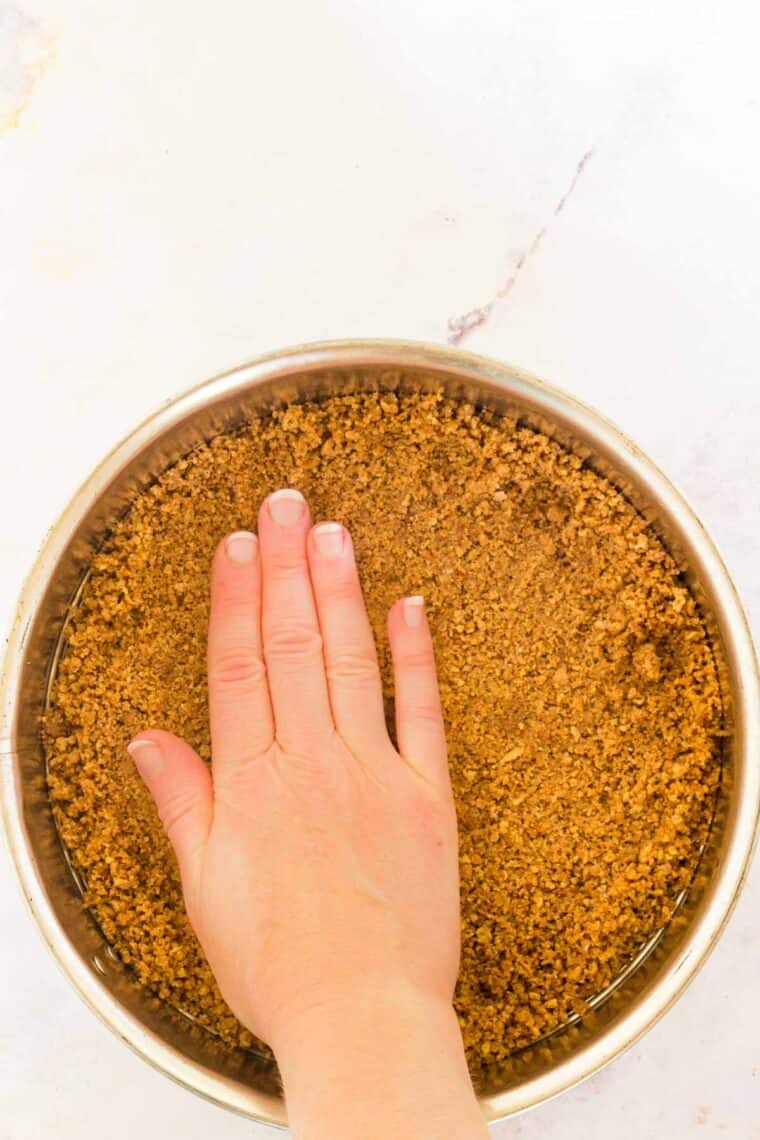 A hand presses gluten-free graham cracker crust into the bottom of a springform pan.