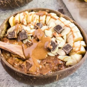 A brown bowl of chocolate oatmeal topped with almond butter, sliced almonds, chocolate chunks, and a sliced banana.