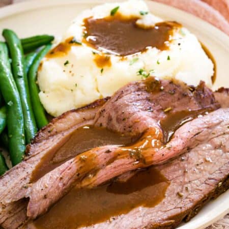 Slices of bottom round roast served on a plate with green beans and mashed potatoes, covered in gravy.