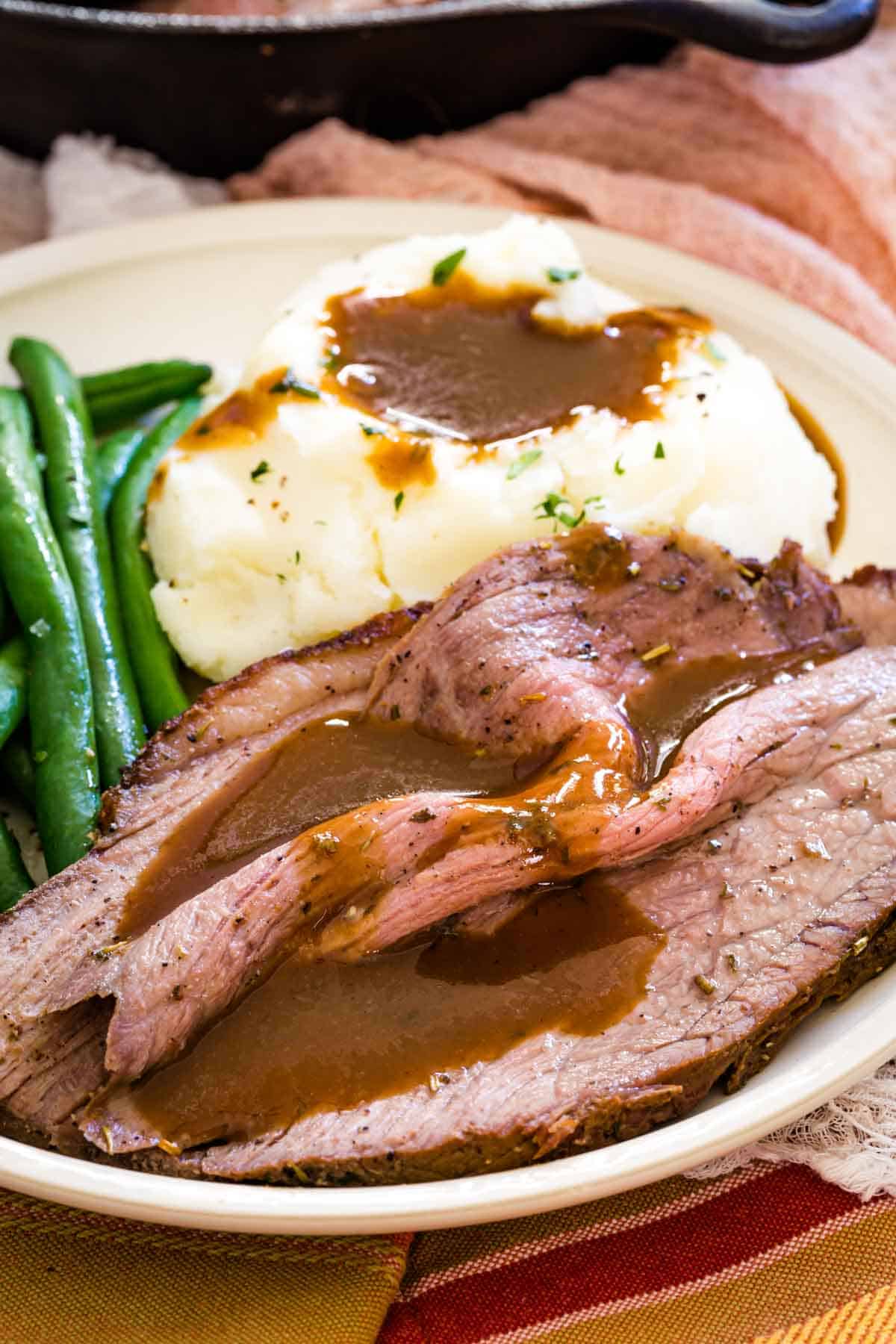 Slices of bottom round roast served on a plate with green beans and mashed potatoes, covered in gravy.