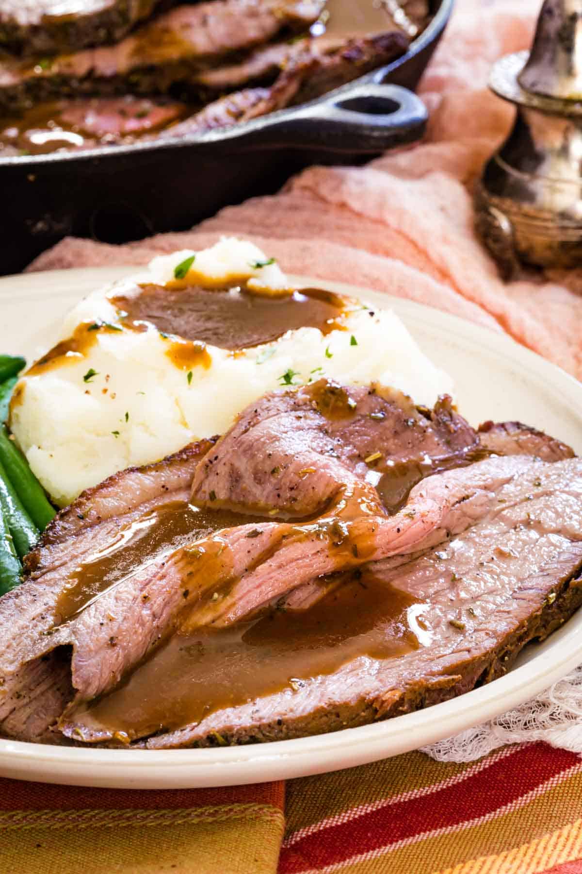 Slices of bottom round roast served on a plate with green beans and mashed potatoes, covered in gravy.