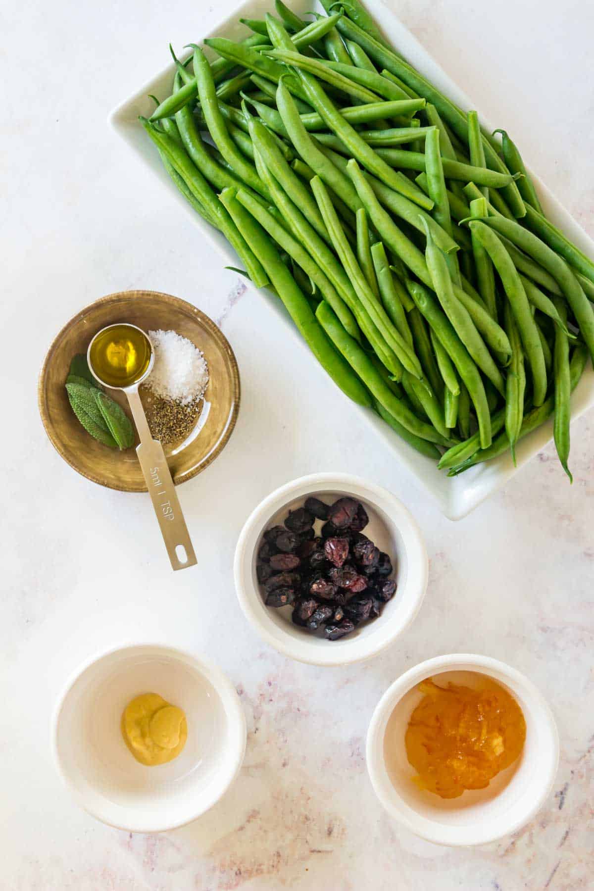 All of the ingredients to make orange glazed green beans with dried cranberries in bowls on a marble countertop.
