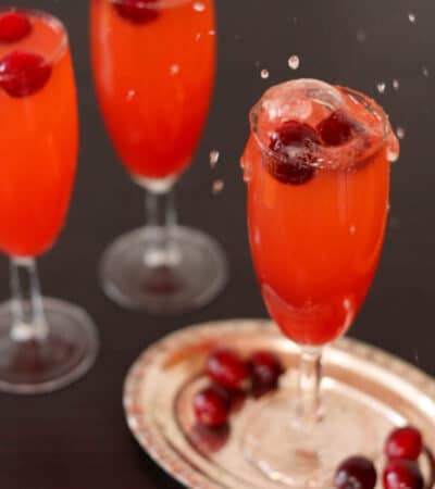 Cranberries splashing into a red mocktail in a chapagne flute with two more of the drinks behind it.