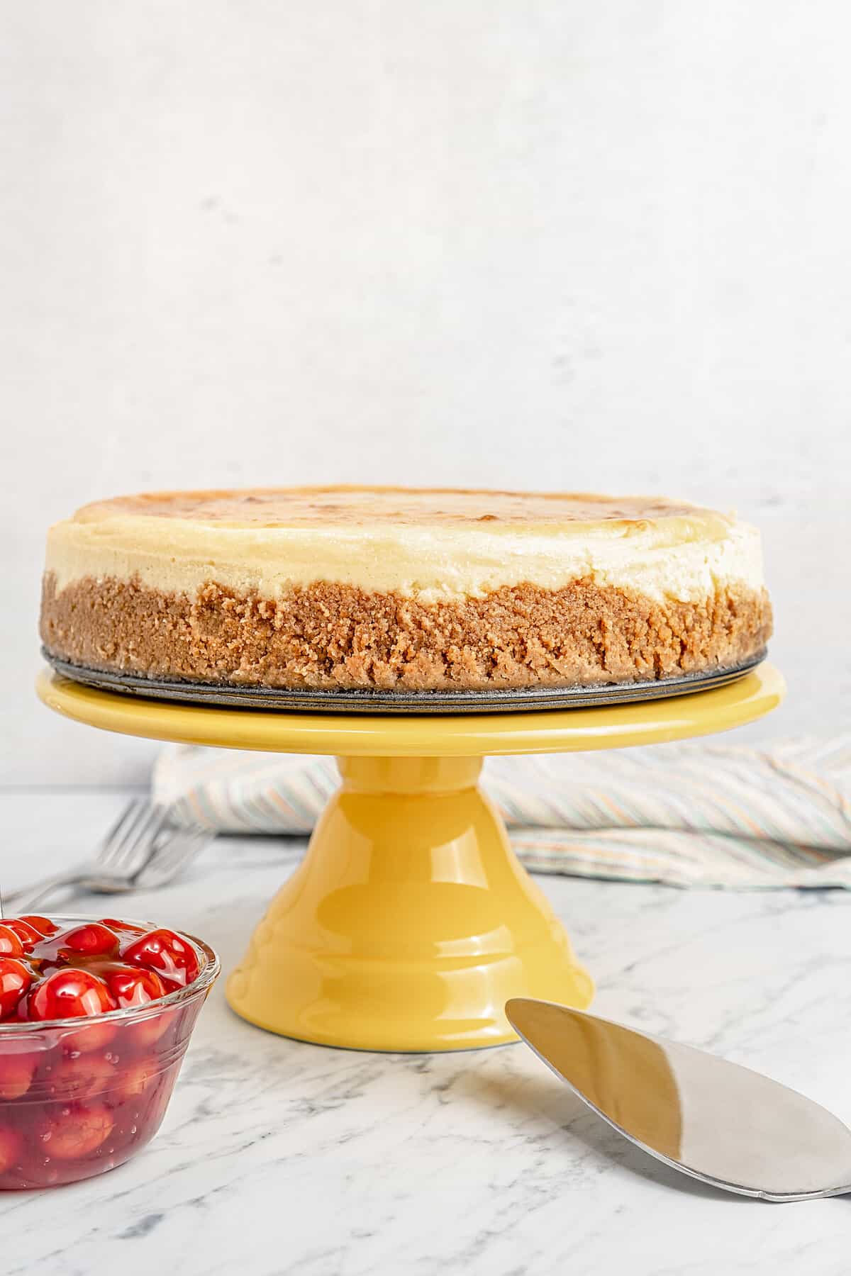 A classic gluten-free cheesecake on a yellow cake stand next to a bowl of cherries.