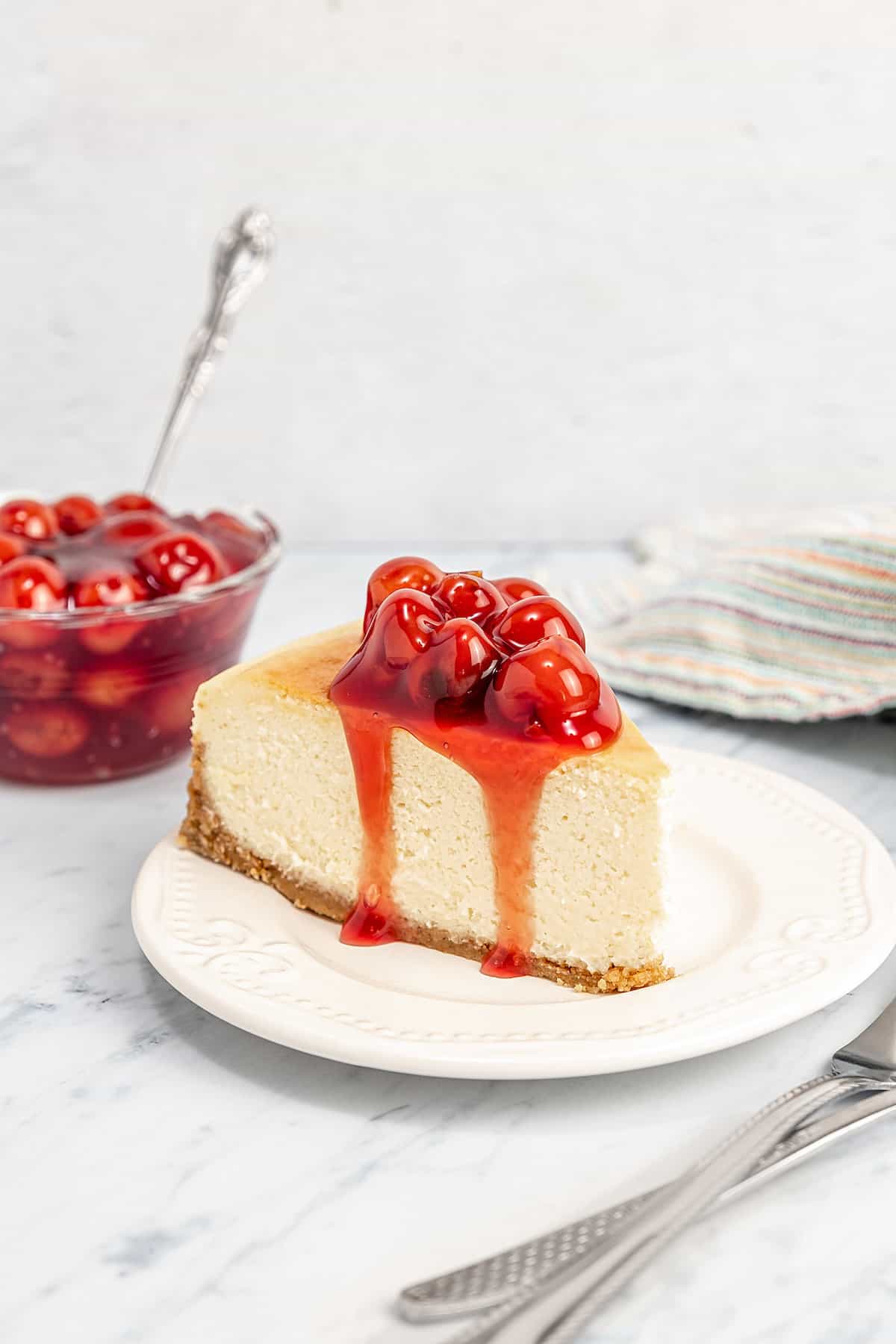 A slice of gluten-free cheesecake topped with cherries on a plate with a bowl of cherries in the background.