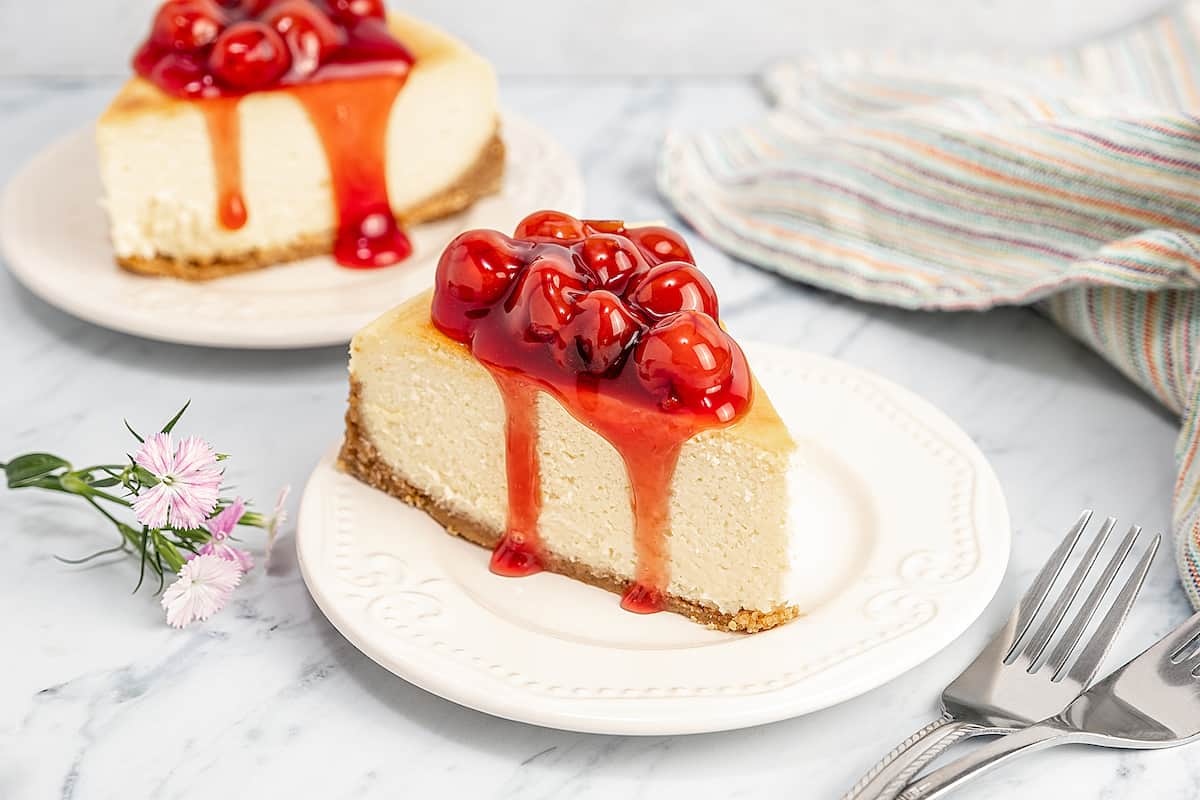 Two slices of gluten-free cheesecake on plates, topped with cherries.